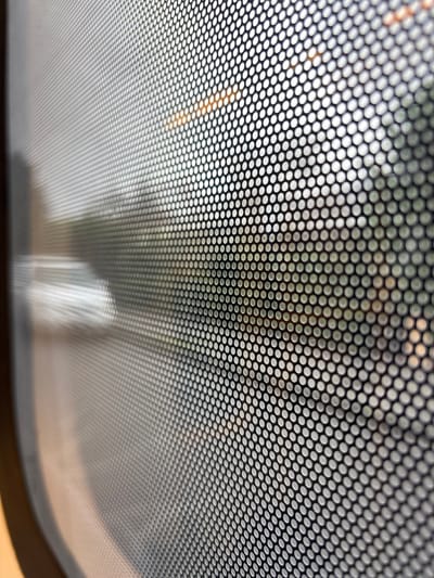 The image is a close-up, angled shot of a perforated screen, likely a window or panel, obscuring the view of the environment outside. The screen has a consistent pattern of small, circular perforations. Through the screen, a blurred scene is visible, suggesting movement or a distant focus. Elements of the outdoor environment include what appear to be trees or foliage with green hues, and hints of orange and yellow lights, possibly from streetlights or buildings. A bright, elongated shape, likely a reflection or a light source, is visible on the left side of the image. The overall impression is of looking through a filtered medium, creating a sense of distance and abstraction. There are no discernible people or specific landmarks to identify Brunswick East, Australia.