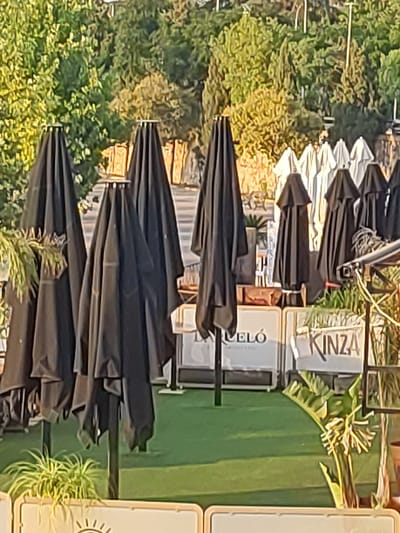 The image shows an outdoor seating area with several closed black umbrellas. In the background, there are several white umbrellas and lush green trees. The scene appears to be a cafe or restaurant patio in Badajoz, Spain, as indicated by the signage visible in the image. One sign reads "KINZA", and another partially visible sign says "ELÓ". The ground is covered in artificial green turf, and there are potted plants with green leaves visible. The overall impression is that of a pleasant outdoor dining space, possibly in the late afternoon given the warm lighting. There are no people visible in the image.
