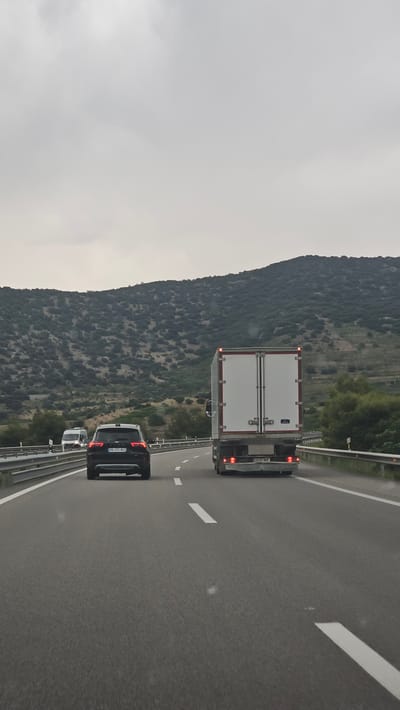 The image depicts a highway scene in El Frasno, Spain, under overcast skies. The camera is positioned behind a black SUV, which is following a white box truck on a multi-lane highway. The truck has a white trailer with red accents and taillights, and its license plate reads "2337 NBP". The black SUV has its brake lights illuminated and its license plate is partially visible, showing "HK 051 AOJ". Further ahead, another white vehicle is visible. The highway is flanked by a steep, vegetated mountainside on the left and a guardrail on the right. The road surface is asphalt and appears wet, suggesting recent rain. The overall atmosphere is somewhat muted due to the cloudy weather, creating a diffused light. The scene captures a moment of travel on a highway, with vehicles moving along the road amidst a natural landscape.