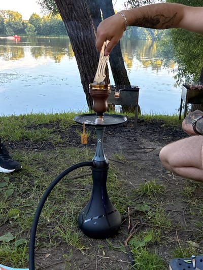 In Il'inskoye, Russia, on a grassy area by a body of water, a person is preparing a hookah. A dark-colored hookah with a grey stem stands prominently in the foreground, its black hose extending to the left. A person's arm, adorned with tattoos and a silver bracelet, is visible above the hookah, holding a tool with tongs to place something in the hookah's bowl. The bowl itself is brown and brass, and a charcoal sits on top. A small, yellow wooden tag hangs from the hookah stem. Two charcoal grills are positioned behind the hookah, one with food cooking on it. The water in the background reflects the trees and sky, and a red boat is visible on the water in the distance. The ground around the hookah is covered in grass and scattered twigs. The lower right corner of the image shows a person's leg and foot wearing a dark blue Croc.