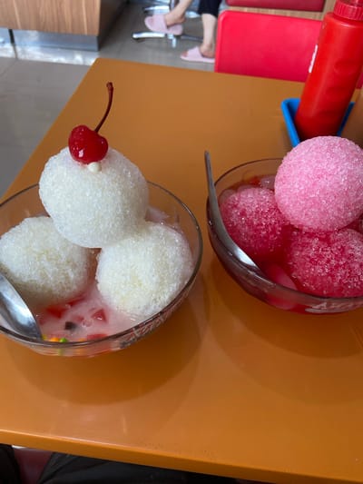 Two bowls of shaved ice desserts sit on an orange table in a restaurant setting, likely in Kudus, Indonesia. The bowl on the left contains three white scoops of shaved ice, topped with a maraschino cherry and a small white pearl. The ice is nestled in a milky liquid with colorful jelly pieces. The bowl on the right holds three pink scoops of shaved ice in a red, syrupy liquid. A red ketchup dispenser is visible in the background, along with part of a red chair and the lower legs and feet of a person wearing pink slippers. The overall environment suggests a casual dining establishment.