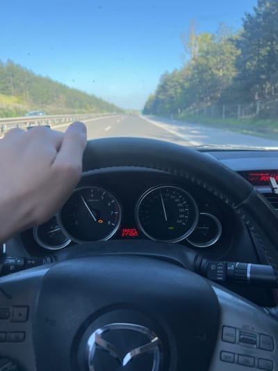 The image shows the interior of a moving car, specifically the dashboard and steering wheel. The odometer reads 260227, with the trip meter showing 21600.3. The speedometer needle is at zero. The engine is at idle. The hand is on the steering wheel, and the Mazda logo is visible. The car is on a road with trees and a blue sky visible through the windshield. There is nothing in the image to confirm that the location is Церово, Bulgaria.