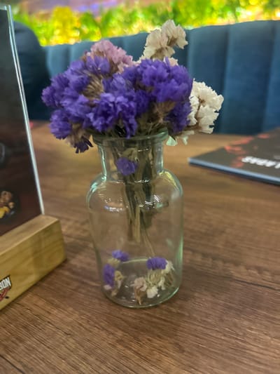 A glass vase containing a bouquet of dried purple and white flowers rests on a wooden table. Part of a menu or booklet with text and images is visible at the bottom of the image. Other menus or reading materials are partially visible to the left of the vase. The table is indoors, and the background shows a wall with a green and yellow pattern to the left and a blue surface behind the vase. The location is La Aurora, Ecuador.
