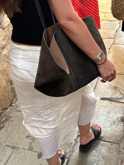 A person is walking on a cobblestone street in Split, Croatia. They are wearing a black tank top, a white skirt, and black slide sandals. The person is carrying a large, dark-colored tote bag and wearing a watch on their left wrist and a ring on their right index finger. The street appears to be narrow and lined with stone buildings, typical of the historic center of Split. The lighting suggests it is daytime, possibly late afternoon due to the shadows.