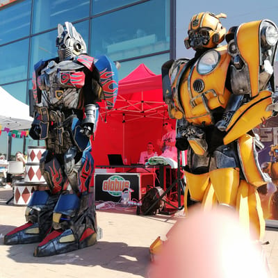 The image depicts an outdoor event in Трубачеевка, Russia, under a clear, bright sky. Two individuals in large, elaborate costumes resembling Optimus Prime and Bumblebee from the Transformers franchise stand on a light-colored paved surface in the foreground.
On the left, the Optimus Prime costume is predominantly dark gray and silver, accented with blue and red panels featuring red flame motifs. Blue lights are visible on its lower legs. The costume faces slightly to the right. On the right, the Bumblebee costume is primarily bright yellow with dark gray components, featuring vehicle-like details such as headlights on its chest and rounded body panels. This costume faces slightly to the left. A large, out-of-focus, reddish object (likely a finger or thumb) partially obscures the lower right corner of the frame.
Behind the costumed figures, a red pop-up tent houses a DJ setup. Inside, one man with a mustache and a white t-shirt is seated behind a black table, operating a laptop. To his right, another man wearing sunglasses and a white t-shirt stands, holding an object in his hand. A black speaker case displaying a "globus" logo in white and green is positioned beneath the table. Cables and a black backpack are visible on the ground near the equipment.
To the far left, a white pop-up tent is visible with colorful triangular bunting strung along its side. Beneath this tent, indistinct figures are partially visible, one seated behind a white chair. Two large, tiered cylindrical props, patterned with red and brown diamonds, stand near the Optimus Prime costume. In the background, a modern building with large, reflective blue-tinted windows reflects the clear sky. A partial poster or banner, displaying the letters "S P E" and an image of Bumblebee, is visible on the far right behind the Bumblebee costume. The sun casts distinct shadows to the left, indicating a midday or early afternoon setting.
