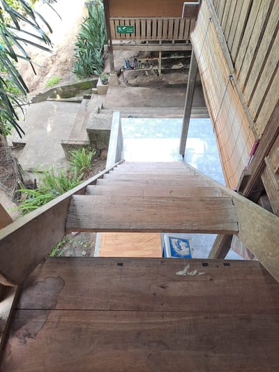 The image provides a top-down perspective of a wooden staircase leading down from a raised wooden structure. The staircase is made of weathered planks. Below the staircase, a tiled floor is visible. To the left of the staircase, a concrete pathway and a lush green fern are present, with a sandy embankment in the background. To the right, a bamboo blind hangs from the wooden structure. A green sign with white text and a pictogram indicating an evacuation route is mounted on a wooden railing of the structure, visible in the upper left portion of the image. Various pairs of shoes are arranged on shelves beneath the raised structure. The scene appears to be an outdoor or semi-outdoor area, possibly part of a dwelling or guesthouse. The location is identified as Geulanggang Baro, Indonesia.
