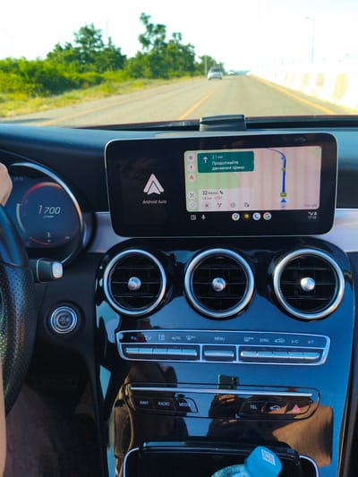 The image shows the interior of a Mercedes-Benz car, likely in Utash, Russia. A person's left hand is visible on the steering wheel, indicating they are driving. The car's dashboard features a digital instrument cluster displaying "1700 x1/min" and a central infotainment screen running Android Auto. The navigation interface on the screen shows a route with instructions in Russian: "3,1 km Продолжайте движение прямо" (Continue straight) and an estimated travel time of "32 мин." The exterior view from the car window reveals a road surrounded by green trees and vegetation, typical of a rural or suburban setting. The weather appears to be bright and sunny. The car's center console with climate controls, media buttons, and other features is also visible.