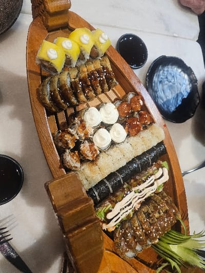 A wooden boat-shaped platter overflows with sushi. A variety of rolls are visible, including those topped with yellow fruit, dark sauce, white cream, and fried tempura with drizzled sauce. Cucumber garnishes the end. Small bowls of dark sauce sit beside the platter. A black bowl rests near the top left corner. Utensils are placed around the table.