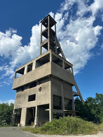 The image depicts an abandoned concrete industrial structure, likely a former mine shaft headframe, under a partly cloudy blue sky. The structure is tall and multi-leveled, with an open framework at the top supporting a concrete tower. The lower sections are more solid concrete, with visible openings that were once windows or doorways, many of which are now empty or damaged. The concrete appears weathered and stained, with some areas showing exposed rebar. Vegetation, including green bushes and white wildflowers, grows around the base of the structure and encroaches onto a gravel path in the foreground. Lush green trees are visible behind and to the right of the building. The overall impression is one of decay and nature reclaiming an industrial site. The location is identified as Rosen, Bulgaria. There are no people or animals visible in the image.