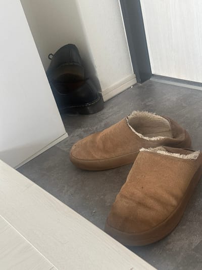 A pair of black shoes sit inside a white shoe cabinet near a gray floor. Next to the cabinet and on the floor is a pair of tan suede slippers, one partially overlapping the other. The right-most slipper is the closest to the right side of the image and at a slight angle.