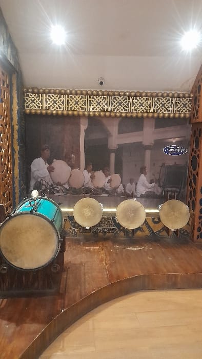 An indoor cultural display in Glantengan, Indonesia, showcases traditional Indonesian percussion instruments. In the foreground, a raised, dark wooden platform features a large, light blue drum with a light-colored drumhead on a wooden stand to the left. To its right, four smaller, circular, light-colored frame drums are arranged in a row on a lower stand, each with a light-colored skin. Behind these instruments, a large sepia-toned mural serves as a backdrop, depicting multiple male figures in traditional attire seated and playing similar drums within a traditional, possibly outdoor, setting with columns. A logo, appearing to read "Mubarok," is visible on the upper right of the mural. Above the mural, a golden decorative frieze with intricate geometric patterns and a row of golden spheres adorns the wall. Ornate wooden panels with cutout patterns flank the display area. The white ceiling features two bright overhead lights and a single white security camera (CCTV) mounted centrally. The floor in front of the platform is a lighter, smooth material, possibly wood or tile. The scene is static, with no active people or direct interactions depicted.