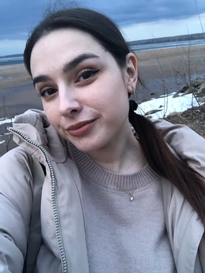 A woman is taking a selfie outdoors. She has dark hair pulled back into a ponytail. She is wearing a light gray sweater under a beige puffer jacket. She has an ear cuff on her left ear. A thin gold necklace with a small pendant is visible. The background appears to be a body of water and land, likely near a shore or riverbank. This does not appear to be in the city of Kizhi, Russia.