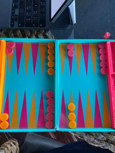 The image displays a vibrant backgammon board, an iPad with a keyboard, and two dice, suggesting a game in progress or about to begin. No people are visible.
The central subject is a colorful backgammon board featuring a teal playing surface with alternating pink and orange triangular points. Pink checkers are arranged with five on the top left point, two on the top left bar, and five on the bottom right point. Orange checkers are positioned with three on the top right point, five on the bottom left point, and two on the bottom right bar. Two red dice are seen near the top left edge of the board, displaying faces with numbers such as 1, 3, 4, and 6.
To the left of the backgammon set, an open iPad rests in a black keyboard case. The iPad's screen is white, and portions of the QWERTY keyboard are visible, including keys labeled "option" and "cmd". The scene is set on a dark, flat surface, with woven, brownish material visible to the right and partly beneath the objects. The lighting is diffuse, making the time of day indeterminate. While the location context is Mto Wa Mbu, Tanzania, there are no specific visual cues within the image to confirm this.