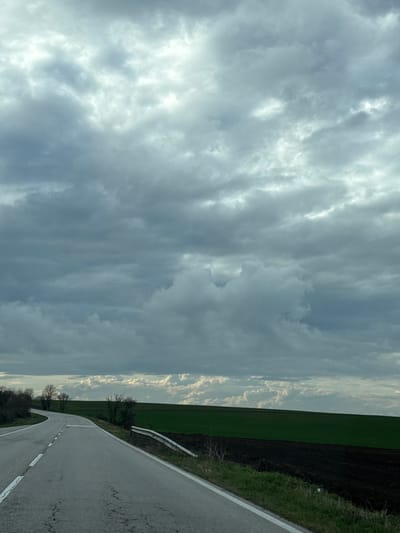 The image shows a view from a road in what could be Bogorovo, Bulgaria. To the left is a sky filled with grey, striated clouds. To the right of the sky is a vertical strip of green farmland bordered by dark soil. A grey asphalt road with a white dividing line runs diagonally across the bottom right, next to the farmland. The road curves slightly into the distance. Guardrails are visible between the farmland and the road. Bare trees are visible along the edge of the field.