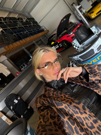 An indoor image depicts a blonde female in a garage or workshop setting located in Meuspath, Germany. The female, positioned in the foreground, wears rimless glasses with light brown tinted lenses, a black zip-up top, and a brown and black leopard print jacket. Tattoos are visible on her neck, and a silver-colored stud earring is present on her left earlobe. Her left hand, adorned with multiple rings and light-colored manicured fingernails, is raised to her chin area. She is looking directly towards the camera.
In the mid-ground, a red performance car with its hood open is visible, featuring yellow brake calipers and black and white sponsor decals on its side. To the right of the red car, a racing helmet with black, white, blue, and yellow graphics, including "RAVEN" and "REHAU" text, rests on a surface. Further in the background, a yellow sports car is partially visible. To the left of the scene, a large metal shelving unit holds numerous stacks of black car tires. The floor is concrete. Additional objects include a black travel bag with white straps and an Amazon-branded cardboard box on the floor to the left of the female. A dark grey and black striped wall panel is visible behind the female on the left side of the frame.