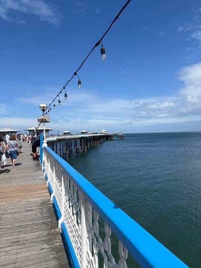 The image shows a view from Llandudno Pier in Llandudno, United Kingdom. The pier extends into the sea, with a blue and white railing visible along the right side of the frame. A string of lights hangs diagonally across the upper left of the image. People are visible walking and sitting along the pier. The sea is visible on the right, and the sky with scattered clouds is on the left.