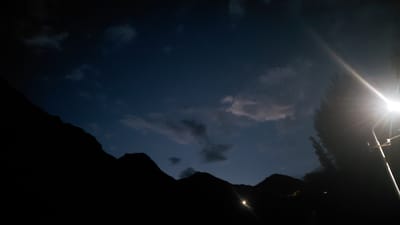 The image depicts a silhouetted mountain range against a deep blue twilight sky. Wispy clouds are scattered across the sky, illuminated by a bright, intense light source on the right side of the frame. This light source appears to be a streetlamp or a powerful artificial light, casting a strong glare and lens flare that dominates the upper right quadrant. A tall, dark silhouette, likely a tree, is positioned between the light and the mountains.

The lower portion of the image is filled with the dark silhouettes of several mountain peaks and ridges, their contours sharply defined against the fading light. In the valley between some of the peaks, there are faint, small points of light, suggesting human habitation or artificial lighting in the distance.

The time of day is clearly dusk or early night, transitioning from daylight to darkness. The atmosphere is calm and serene, with a sense of quietude, though the powerful artificial light introduces a stark contrast. The location is identified as Ak-Terek, Kyrgyzstan, which, combined with the mountainous landscape, suggests a rural or remote setting. There is no visible text in the image.
