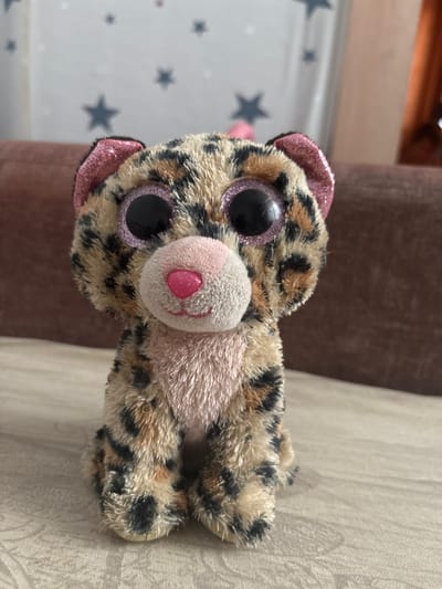 A plush toy leopard with large, sparkly pink eyes and a pink nose sits on a textured, light-colored surface, possibly a sofa or bed. The toy has a tan and black spotted fur pattern, with pink accents on its ears. The background is blurred, showing a brown upholstered surface and a wall with a faint pattern of dark blue stars. The lighting suggests an indoor environment. The scene appears to be in a residential setting, possibly in the city of Клейнікаўскі сельскі Савет, Belarus. There are no people or actions visible in the image.