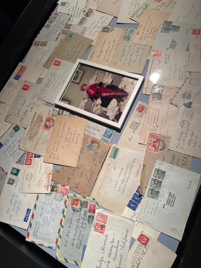 The image displays a collection of envelopes, many addressed to Marilyn Monroe at 20th Century Fox Studios in Beverly Hills, California. Several envelopes are stamped with postmarks from various locations, including U.S.A., Germany, and possibly other international origins. Some envelopes are open, revealing handwritten correspondence. A framed photograph of Marilyn Monroe is placed among the envelopes, showing her in a red garment surrounded by more papers. The collection is laid out on a flat surface, possibly a display in a museum or archive. The background suggests a dark, enclosed environment, consistent with an indoor exhibition space. The overall impression is of a curated display of personal correspondence related to the iconic actress. While the image itself does not provide specific details about a location in Wattens, Austria, this collection of correspondence is housed within the Swarovski Kristallwelten in Wattens, Austria, as part of a special exhibition.