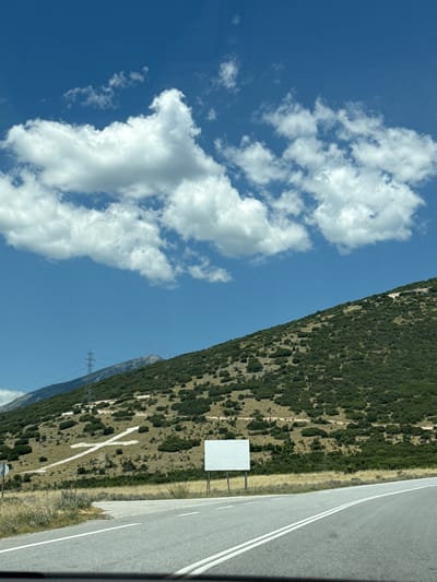 The image depicts a sunny day with a clear blue sky and scattered white clouds above a rugged, green hillside. A white cross, formed by clearing vegetation, is visible on the slope of the hill. A road curves through the foreground, with white lines marking its lanes. Adjacent to the road, a blank white billboard stands on poles. In the background, a power transmission tower is visible against a mountain range. The scene appears to be located in the Municipal Unit of Prosotsani, Greece, suggested by the visual context of the landscape.