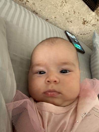 A close-up, eye-level selfie shows a baby, likely female due to the pink ruffled garment with sequins, looking directly at the camera. The baby has light skin, dark eyes, and plump cheeks. The background consists of a striped grey fabric, possibly a blanket or bedding, and a textured brown surface with dark speckles. A smartphone is visible in the upper right corner, displaying a colorful screen. The image appears to be taken indoors, in a residential setting. There is no indication of the location being the city of Serebryakovka, Russia.