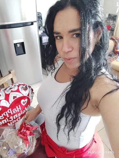 The image shows a person, appearing to be an adult with long, dark, wavy hair, looking towards the camera. The image is oriented sideways, with the person's head on the right side and feet towards the left.

The person is wearing a plain white sleeveless top and red bottoms, possibly trousers or a skirt, featuring a prominent red bow tied at the waist. Their left arm is visible, holding a red and white foil balloon and a gift wrapped in clear cellophane.

The balloon is heart-patterned and appears to have text in white, with "Happy Valentine's" partially visible, suggesting it's a Valentine's Day balloon. The gift, wrapped in clear plastic with a red ribbon, seems to contain a light-colored, plush item, possibly a teddy bear or similar soft toy, accompanied by other contents that are not clearly identifiable.

The background includes a large, metallic refrigerator with a dark dispenser panel on its front, positioned to the right of the person's head. Above the gift on the left, a wooden structure, possibly part of furniture or shelving, is visible. The floor appears to be a light, possibly tiled, surface.

In the bottom left corner of the image, a date and time stamp reads "14/02/2023 11:53", confirming the image was captured on Valentine's Day morning. The person is looking directly at the camera with a neutral expression, posing with the celebratory items, suggesting they have either received or are presenting the gift and balloon.