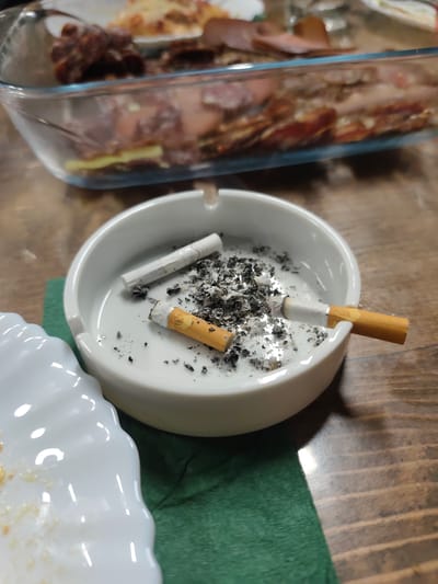 The image shows an ashtray filled with cigarette butts and ashes, resting on a green napkin atop a wooden table. A partially visible white plate is also present. In the background, there is a rectangular glass dish containing a dish with layered ingredients, including meat. The location is indicated to be Mosomishte, Bulgaria.
