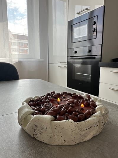 The image depicts an indoor scene, likely a kitchen, with a large, decorative candle in the foreground. The candle is housed in a rough-textured, off-white bowl and is filled with dark brown, bean-shaped objects, possibly resembling coffee beans, embedded in a reddish-brown wax. Two small flames are visible, indicating the candle is lit.

Behind the candle, a grey countertop is visible, leading to kitchen cabinetry. To the left, a portion of a dark upholstered chair is present. In the background, a window with sheer white curtains reveals a glimpse of an apartment building with red brick accents and a cloudy sky. Adjacent to the window, a modern black built-in oven and microwave unit is installed in white cabinetry. The overall environment appears to be a domestic setting. The image does not provide any information about the city of Malye Ust'ya, Russia.