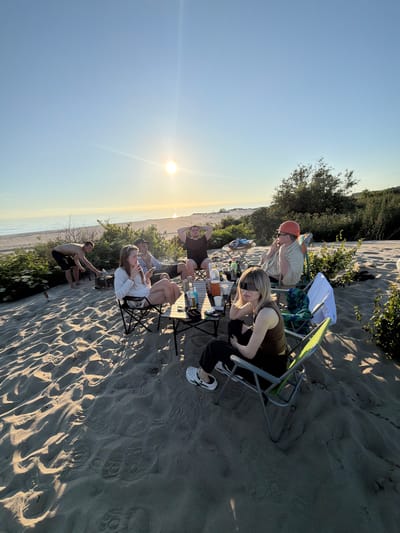 A group of six people are gathered on a sandy beach near the sea, likely in Povorovo, Russia, during sunset. The sun is low in the sky, casting a warm glow and long shadows across the sand. The group appears to be enjoying a casual outdoor gathering, with some seated in camping chairs around a low table. On the table, there are bottles of drinks, cups, and what looks like food. One person is tending to a small grill or fire pit. The environment is natural, with sand dunes, sparse vegetation, and the sea stretching to the horizon. The overall mood is relaxed and social.