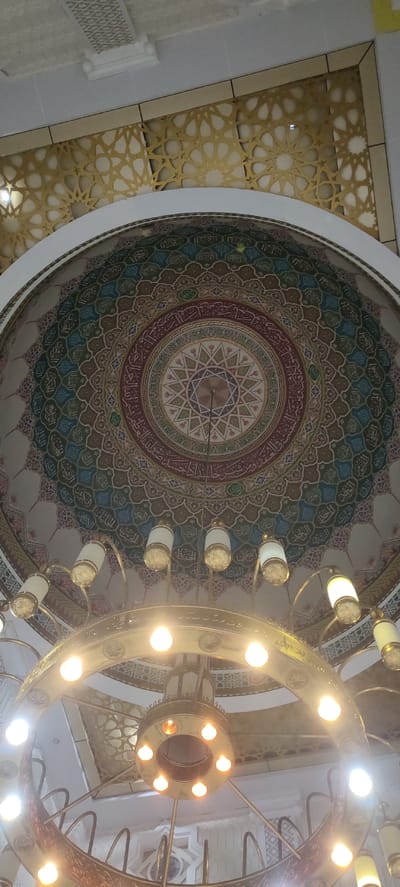 The image captures an ornate dome interior of what appears to be a mosque in Bireuenmeunasahdayah, Indonesia. The dome is elaborately decorated with intricate geometric patterns and calligraphy in vibrant colors, including shades of blue, green, red, and gold. A large, multi-tiered chandelier with numerous light fixtures hangs prominently in the foreground, illuminating the space. The chandelier has a metallic finish, likely brass or gold-toned, and features decorative carvings. Above the dome, a section of the ceiling is visible, adorned with geometric latticework in a gold-colored material. The overall impression is one of grandeur and religious artistry. The lighting suggests it could be daytime or evening, with the artificial lights of the chandelier playing a significant role in the illumination. There is no visible activity or people in the frame, focusing solely on the architectural and decorative elements of the space. The calligraphy on the dome, if decipherable, could provide further context about the specific verses or dedications, but the text itself is not clearly legible in this image.