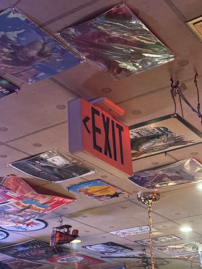 The image shows a ceiling adorned with various posters and an illuminated red exit sign. The posters appear to be advertisements or decorative items, featuring imagery of cityscapes, wildlife, and possibly historical figures. Some posters are covered in a clear plastic to protect them. A small, old-fashioned toy vehicle is suspended from the ceiling by a hook. A decorative chandelier hangs in the center of the frame. The ceiling itself is constructed from modular tiles. The overall impression is that of a quirky or themed establishment. The location in Adachi, Japan, is not evident from the visual information present in the image.