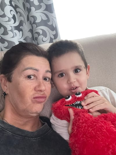 The image features two people, a child and an adult, lying down on a couch. The child is positioned above the adult, with their head close to the adult's. The child is holding a red, plush toy with googly eyes and sharp, black and white teeth. The adult is looking towards the camera with an open-mouthed expression, appearing to be making a "fish face." They are wearing a dark-colored top and a necklace. The child is looking towards the camera with wide eyes, and appears to be smiling slightly. The setting seems to be indoors, with a couch as the primary visible furniture. To the left of the image, patterned curtains are visible. The overall mood of the image appears to be playful and affectionate, suggesting a moment of shared enjoyment between the child and the adult. The lighting suggests it could be daytime. No text is visible in the image.