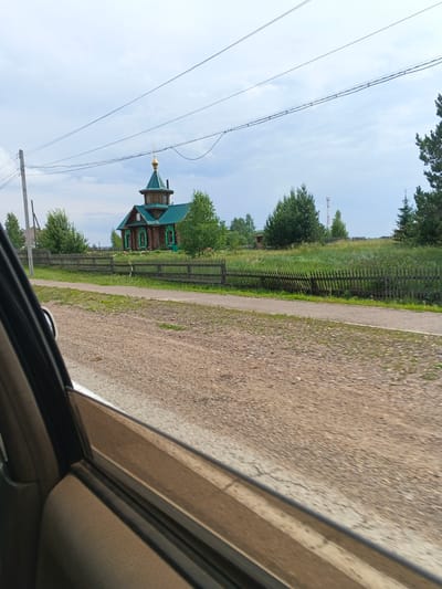 An outdoor, eye-level, medium shot captures a rustic wooden church with a green roof and golden dome, situated in the village of Mokrushinskoye, Russia. The church is flanked by lush green trees and a weathered wooden fence, with a dirt road and a paved walkway in the foreground. The sky above is overcast with scattered clouds. The perspective is from inside a vehicle, with the car's interior and door visible on the left side of the frame.