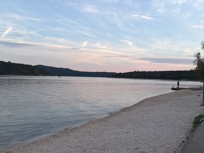 The image depicts a coastal scene at dusk in Banići, Croatia. The foreground features a sparsely vegetated, pebble-covered beach bordering a calm body of water. In the mid-ground, a solitary figure stands on a small concrete pier extending into the water. The background is dominated by a low, dark, tree-covered hill range that extends across the horizon. The sky above is a soft blend of blue and pastel hues, with wispy clouds streaking across it, indicating the transition from day to night. The water reflects the muted colors of the sky and surrounding landscape. A small yellow boat is visible in the distance on the left side of the frame.