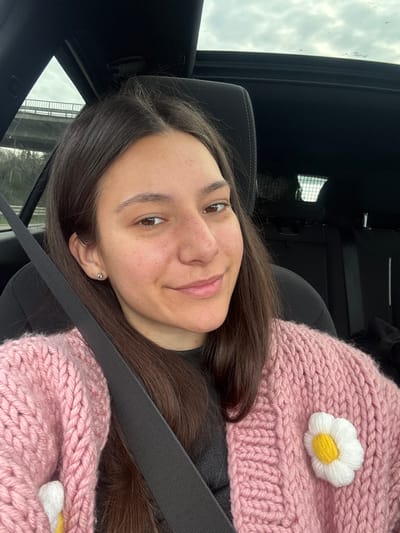 A young woman is seated inside a vehicle, likely a car, wearing a pink, chunky-knit scarf with a white and yellow flower embellishment. She has dark hair and is wearing a seatbelt. She is looking directly at the camera with a slight smile. The vehicle's interior is visible, including the roof and seat. The background seen through the windows is indistinct, but appears to be outdoors with cloudy skies. There is no indication of Kipra, Bulgaria, within the visual content of the image.