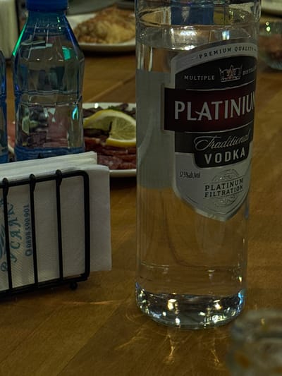 The image shows a bottle of "Platinum" traditional vodka resting on a wooden surface, likely a table. The bottle is positioned horizontally. A blue bottle of water and a napkin holder filled with white napkins are next to it. A plate with food items, including a lemon slice and some cold cuts, is partially visible in the background. A telephone number (0898 590 901) is visible on one of the napkins in the napkin holder. Based on the OCR, this is an advertisement for a place named "CA", which might be a restaurant or bar. The details on the label indicate that the "Platinum" vodka is a multiple distilled beverage with "Platinum Filtration" and a 37.5% alcohol by volume content. The location information provided (Zlati Voyvoda, Bulgaria) implies this scene is set in Bulgaria. The overall scene appears to be set in an indoor dining environment.