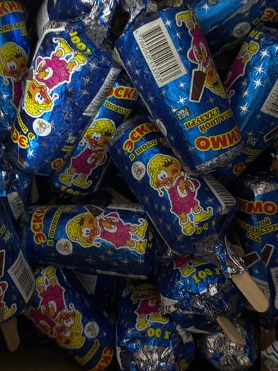 The image shows a close-up, overhead view of a pile of individually wrapped ice cream bars. The wrappers are predominantly blue with shiny metallic accents, featuring cartoon characters of children, stars, and text in a Cyrillic script. Some of the text visible includes "ЭСК" (Esk) and "ШОКОЛАД" (Shokolad), suggesting "ice cream" and "chocolate." The ice cream bars are cylindrical and appear to be on sticks, with the wooden stick ends visible on some. The image is consistent with a retail display or storage of packaged frozen treats. There is no indication of location within the city of Novopokrovka, Kyrgyzstan, or any specific environmental context beyond the pile of ice cream.