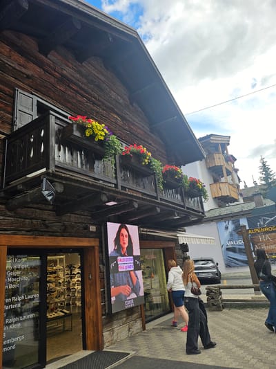 In Livigno, Italy, an outdoor scene features a traditional wooden building with a balcony adorned with flower boxes filled with red, yellow, and purple flowers. A large digital screen is affixed to the lower part of the building, displaying an advertisement for "EDER SHOES" with a woman in a denim jacket and the text "SALDI ESTIVI Fino al 40%". To the right of the digital screen, a sidewalk runs alongside the building, and beyond that, a road is visible. A silver car is parked on the road. Several people are present: two women are walking away from the storefront, one in a white hoodie and blue shorts, the other in a white tank top and dark wide-leg pants, carrying a purse. Another woman in dark jeans and a black top walks away from the scene. Across the street, a sign for "ALPENROSE HA HOTEL" is visible. The environment suggests a commercial area in a mountainous region, with a cloudy sky above. The architecture of the wooden building is typical of alpine regions.