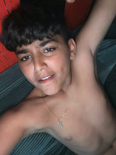 A young individual, appearing to be male, is depicted in a close-up, high-angle portrait, lying on their back with their head slightly tilted to the left. They possess dark, short, curly hair and dark eyes, looking directly at the camera with a gentle, relaxed smile that shows their front teeth. The subject is shirtless, revealing a smooth, medium-toned complexion. A thin gold chain with a small, simple cross pendant rests on their chest.
The person's arms are outstretched and rest on the surfaces beside them. The background consists of two distinct textures: a red fabric with subtle patterns beneath their head, and a dark teal or greyish-blue ribbed or corrugated fabric under their torso and right arm. The lighting is dim, suggesting an indoor setting with low light conditions, possibly evening. The overall emotion conveyed by the subject is calm and inviting. There is no visible text in the image. The location context provided for this image is Topolchane, Bulgaria.