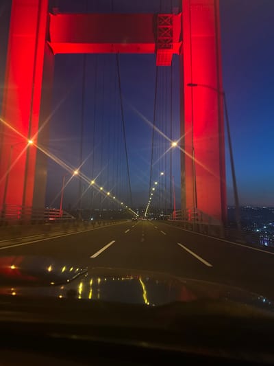 The image depicts a nighttime view from the Yavuz Sultan Selim Bridge in Dilovası, Turkey. The view is directed along the roadway of the bridge, which is divided by white lane markings. Rows of lights illuminate the bridge. The bridge's support structures are painted red and are prominently featured on the left side of the frame. Cables stretch upwards from the support structure to the top of the image. The sky is a gradient of blue to darker shades. In the distance, city lights are visible. The right side of the image shows the interior of a vehicle, suggesting the picture was taken from inside the car. The reflections of the bridge lights are visible on the car's exterior surface.