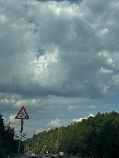 The image depicts a roadside view on a cloudy day, with a highway curving through a densely forested, hilly terrain. In the foreground, a traffic sign warns of slippery roads, indicated by a car skidding on a curve. Below this, another sign indicates conditions of rain and snow. Several vehicles are visible on the road, including a white car and two vans. The sky is filled with dramatic, heavy clouds, with patches of blue sky peeking through. The vegetation is lush and green, typical of a mountainous or hilly region. The overall impression is one of a scenic, possibly remote, stretch of road. The location is identified as Dolno Varshilo, Bulgaria.