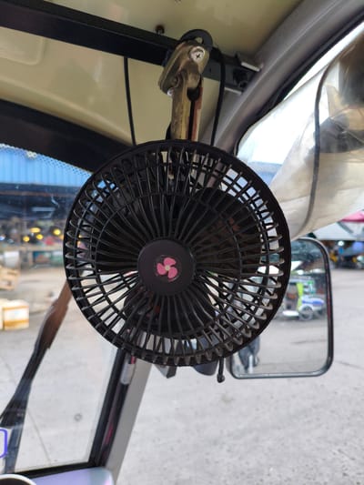 A black, circular electric fan with a pink fan emblem in the center is mounted to the interior ceiling of a vehicle. The fan is positioned in the foreground, obscuring much of the vehicle's interior and the view outside. The vehicle appears to be a tricycle or similar open-air transport, suggested by the presence of a large side mirror visible to the right. The background shows a blurred outdoor scene with buildings, what appear to be market stalls, and other vehicles, indicating a public or commercial area. The overall impression is that of a warm day, given the need for a fan. The time of day is indeterminate, but the lighting suggests daytime. No text is visible in the image.