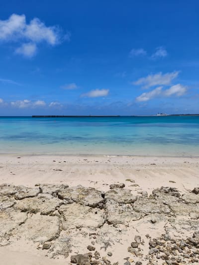 This is a photograph of a beach in Miyakojima, Japan, likely during daytime under a clear sky. In the foreground, there is a sandy beach with some scattered rocks and debris. The sand appears light in color. Beyond the beach, the ocean stretches out, with the water transitioning through shades of turquoise and blue, indicating varying depths.
In the middle ground, a long, dark structure, possibly a breakwater or pier, extends across the horizon. There are some small structures or lights visible on this structure. Further in the distance, a white ship is visible on the water.
The sky above is a vibrant blue, dotted with a few fluffy white clouds, suggesting fair weather. The overall scene depicts a serene coastal environment.