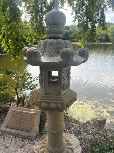This is a photograph of a Japanese stone lantern situated beside a tranquil pond with lush green trees and foliage in the background. The lantern is intricately carved and appears to be made of weathered stone. In front of the lantern, a bronze plaque is mounted on a stone base, bearing an inscription that reads: "THE ASIAN-AMERICAN FEDERATION OF FLORIDA" and "THE JAPAN-AMERICA SOCIETY OF SOUTH FLORIDA RESPECTFULLY DEDICATE THIS LANTERN TO THE MEMORY OF COL. ELLISON SHOJI ONIZUKA AND HIS CHALLENGER CREWMATES JAN. 25, 1987 IN COOPERATION WITH AMERICAN AIRLINES WASTE MANAGEMENT OF FLORIDA". The pond's surface is partially covered with green aquatic plants, and the water reflects the overcast sky and surrounding greenery. The overall scene suggests a serene and memorial garden setting, likely within the Morikami Museum and Japanese Gardens in Delray Beach, Florida, given the inscription referring to the Challenger astronauts and the presence of a Japanese-style lantern.