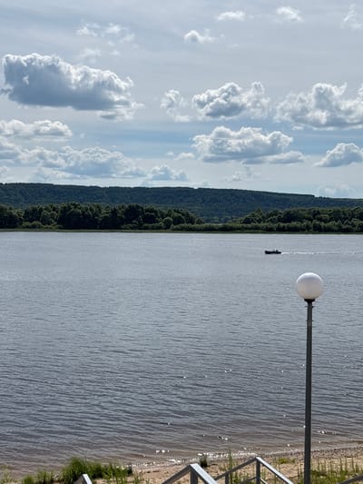 This is a vertically oriented, eye-level shot of a body of water, likely a river, on a cloudy day. The scene is divided into distinct sections. On the left, a vast expanse of sky is filled with dramatic, fluffy white cumulus clouds against a pale blue backdrop. The clouds create interesting shadows and shapes, suggesting movement and depth.
Adjacent to the sky, on the left side of the image, is a dense, dark green forest that slopes down to the water's edge. The trees are tightly packed, creating a textured wall of foliage that contrasts with the lighter tones of the sky and water.
The majority of the image is dominated by the water's surface, which is rippled and shows glints of sunlight reflecting off its surface, indicating it's likely daytime. In the middle distance on the water, a small, dark silhouette of a boat or a person in a kayak can be seen, creating a sense of scale within the expansive environment. Closer to the right side of the frame, a tall, modern lamp post with a spherical white light fixture stands at the edge of the water, suggesting a developed area like a park or promenade. Further to the right, a sandy embankment with some sparse green vegetation is visible, and a metal railing can be seen in the foreground, implying a man-made structure bordering the natural landscape.
The overall mood is one of serene natural beauty with a touch of human presence. The weather appears to be partly sunny with periods of cloud cover. The location cues, such as the dense forest and the wide body of water, along with the context provided, point to a location in Zvenigovo, Russia. No visible text is present in the image.