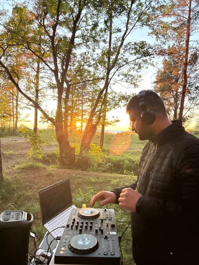 A man wearing headphones and a black quilted jacket is standing in front of DJ equipment outdoors. The DJ setup includes a laptop, a DJ controller, and a speaker. The man is looking down at the DJ controller and his hands are positioned over it, suggesting he is actively mixing music. The scene is set in a wooded area with trees and tall grass. The sun is setting, casting a warm golden light across the landscape and creating lens flares. In the background, a body of water, possibly the Baltic Sea, is visible along with a faint horizon line. The location is identified as Meremõisa, Estonia. The overall impression is that of an outdoor DJ performance at sunset.