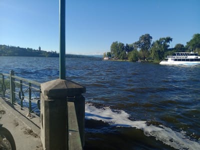 The image presents a scenic view across a wide body of water, likely Lake San Roque in Villa Carlos Paz, Argentina, under a clear blue sky. In the immediate foreground, a concrete walkway runs along the left, featuring a robust concrete pillar and a green metal railing. Part of a vehicle's wheel is visible at the bottom left, and some debris is scattered on the walkway.
The expansive water in the midground is a deep blue, exhibiting a choppy surface with noticeable waves and white foam near the foreground, indicative of windy conditions. On the right side, a large, white multi-deck boat, possibly a tour vessel with the name "ZARIBELLE" faintly visible on its hull, is moored close to a lush, tree-lined shore.
The distant shoreline is characterized by rolling hills blanketed in green foliage, dotted with various buildings. A distinctive, tall, reddish-brown tower-like structure stands out on a distant hillside to the far left. Further along the distant shore, a small pier or dock extends into the water, with a few indistinct figures visible on it.
The sky is a bright, cloudless blue, suggesting a sunny day. Strong shadows cast by the foreground elements confirm it is daytime, likely mid-morning or afternoon. The scene conveys a sense of a picturesque and vibrant waterfront, typical of a popular lakeside destination.