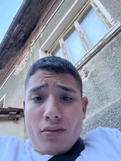 The image is a close-up, low-angle selfie of a young person with dark hair and fair skin. They are looking directly at the camera with a neutral expression. The person is wearing a white t-shirt. In the background, a weathered building with peeling paint and exposed brickwork is visible. A window with a cracked frame and faded paint is also present. A section of a gutter and roof tiles can be seen at the top left of the image. The scene appears to be outdoors in a location that suggests it might be part of General Todorov, Bulgaria, based on the architectural style and evident signs of age and weathering on the building.