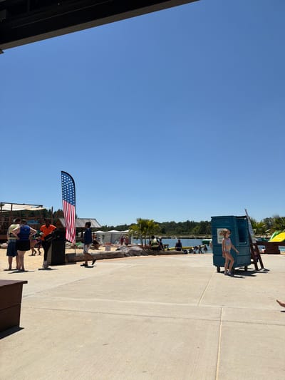 This image depicts a bright, sunny day at an outdoor water park in Wisconsin Dells, USA. In the foreground, a concrete pathway leads towards a sandy beach area where people are enjoying recreational activities. A tall, feather-shaped American flag stands prominently near the pathway. To the right, a young girl in a swimsuit walks past a teal-colored lifeguard stand. In the background, a body of water with several people swimming or floating is visible, with trees and buildings lining the shore. The overall atmosphere suggests a lively and enjoyable summer day at a popular tourist destination.