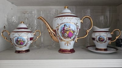A three-piece porcelain tea set, including a teapot, sugar bowl, and creamer with a saucer, is displayed on a wooden shelf. The set is white with gold accents and features a decorative band with a scene of two figures in 18th-century attire. Several clear, cut-glass wine glasses are visible behind the tea set. The shelf is located inside a light-colored wooden cabinet. The scene is likely a domestic interior, possibly a display cabinet in a home.